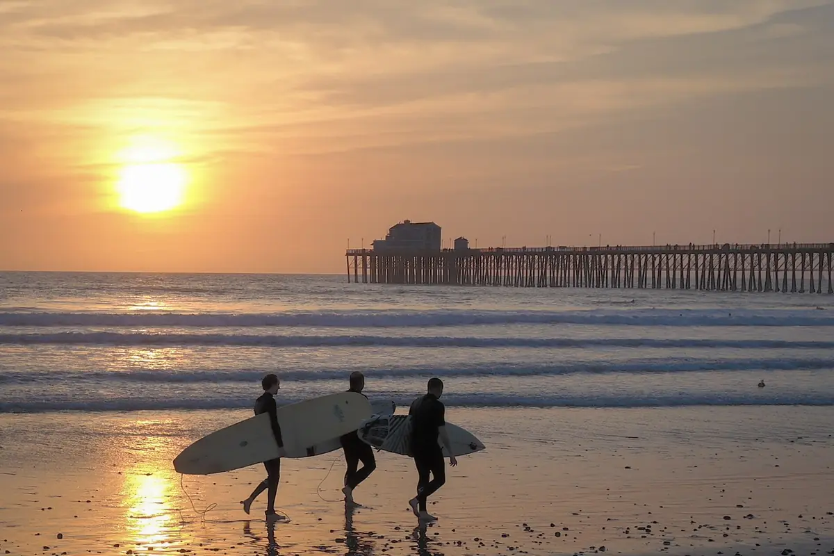 Three surfers carrying boards along the beach at sunset near the Oceanside Pier