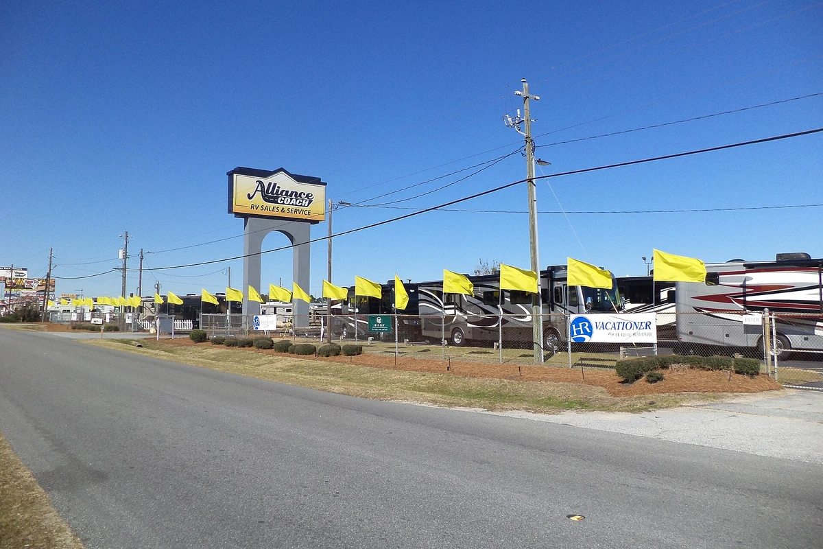 A row of large motorhomes behind a fence at an RV dealer, with yellow flags along the road and a tall dealer sign
