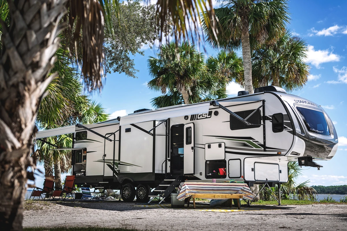 A large white fifth wheel trailer with its awning out and steps down, parked under palm trees beside a lake