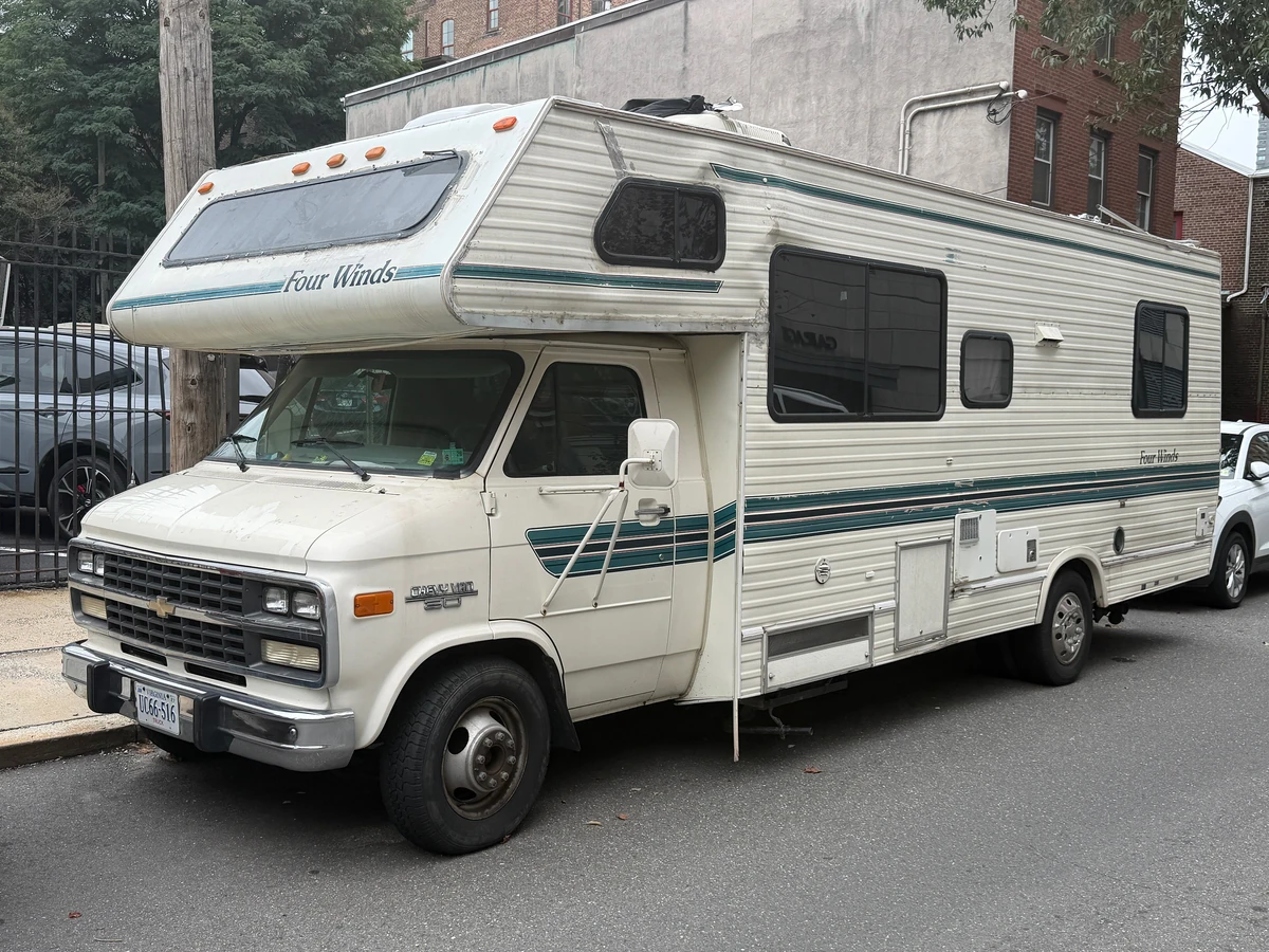 An older cream Four Winds Class C motorhome with teal stripes parked at a city curb, cab over bunk above the driver's seat