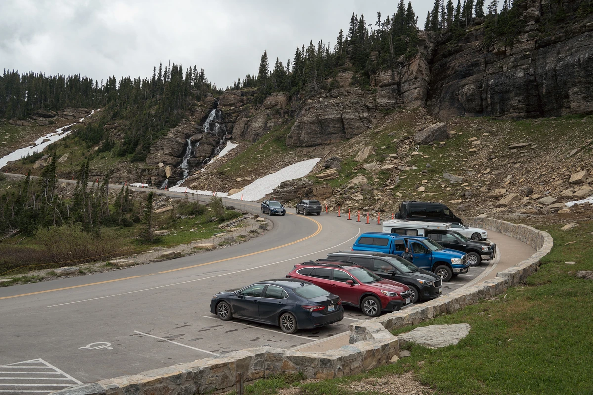 Cars and pickups parked along a stone wall at Oberlin Bend on Going to the Sun Road, with a waterfall and snow on the slope behind