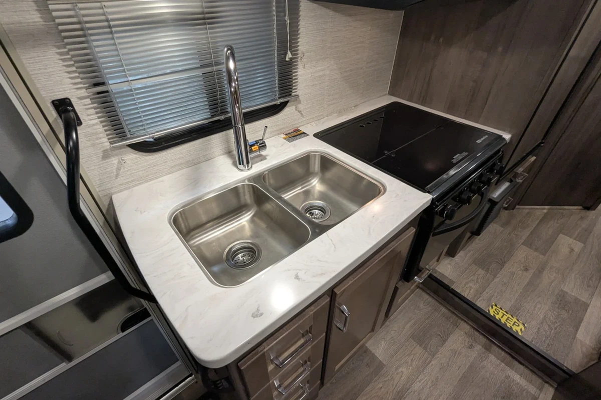 Inside a Class C motorhome galley with a double stainless sink, a black glass two burner cooktop and wood look cabinets