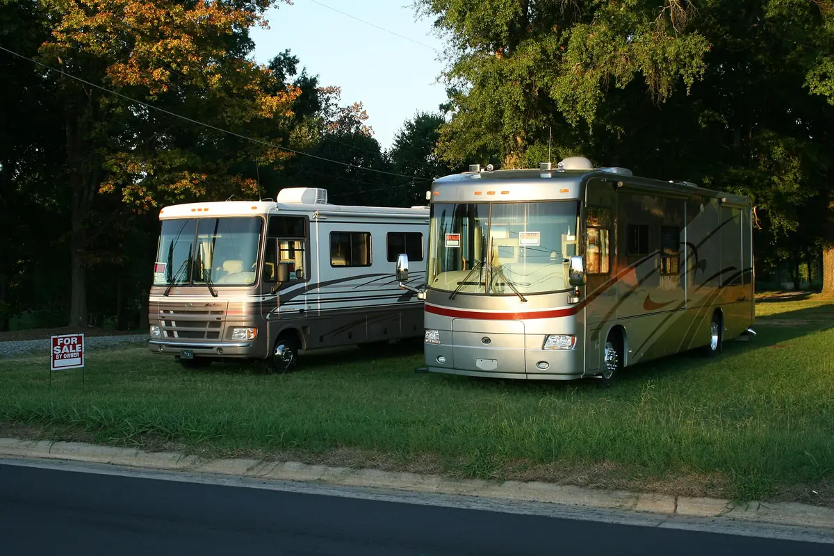 Two Class A motorhomes parked on a grass lot by a street with a for sale by owner sign in front