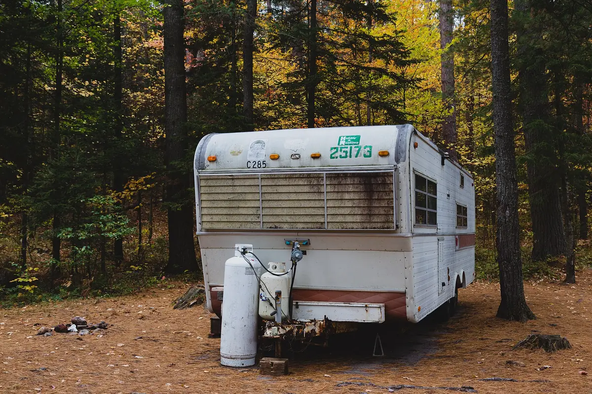 An old white travel trailer parked in autumn woods with dark water stains on its front cap, two propane tanks and a rusty hitch