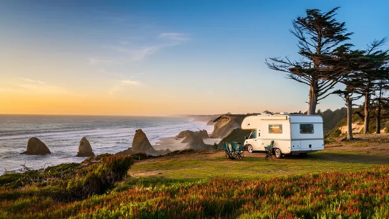A family sitting around a campfire beside a motorhome at golden hour, with pines and a mountain ridge behind them