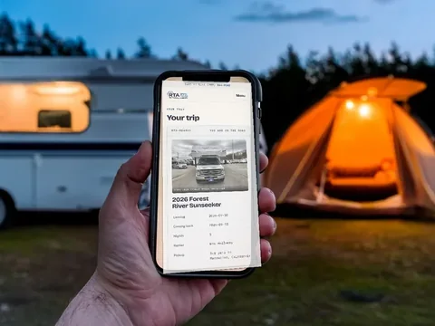 A hand holding a phone at a campsite in the evening with a lit awning behind